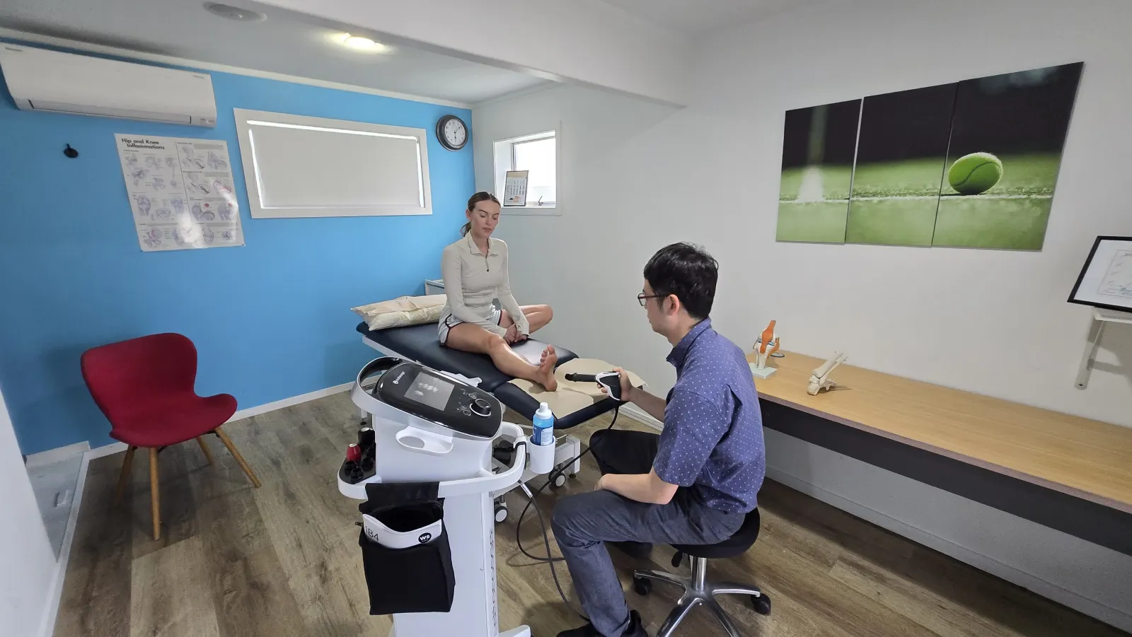 A treatment room at Twins Physio & Podiatry: a clinician treats a patient's heel with a handheld device beside a blue feature wall