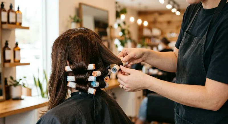 A stylist wrapping sections of long dark hair around colourful perm rods on a client seen from behind, in a warm modern salon